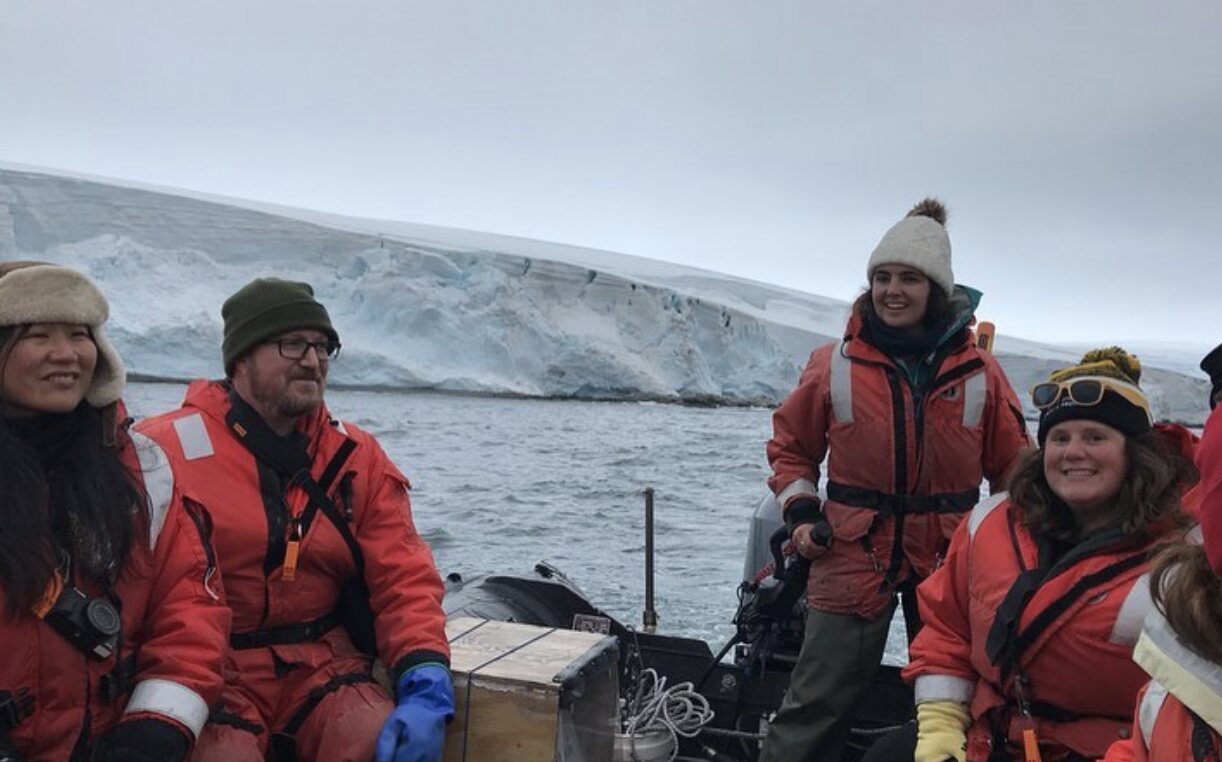 A field team smiling together in a small boat beside an Antarctic glacier