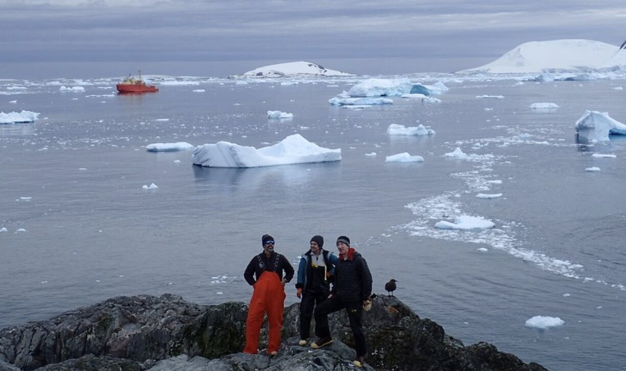 A field team standing above iceberg-filled Antarctic waters with a research vessel in the distance